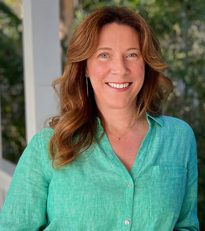 Lisa-Kenchel Lisa Kenchel, founder of Kenchel Interior Design, smiling in a green button-up shirt against a natural outdoor background.