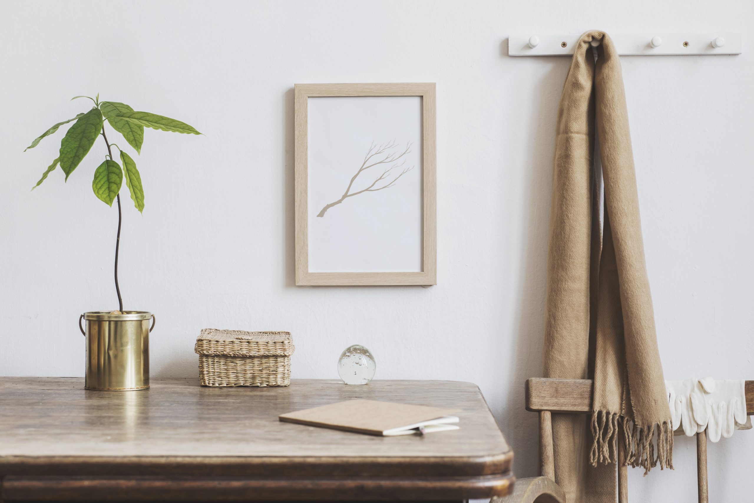 Depositphotos_237628336_XL A minimalist workspace featuring a wooden desk with a small potted plant in a gold container, a wicker storage box, and a notepad with a pen. A framed artwork of a delicate branch hangs on the white wall above, adding a natural and serene touch to the space.